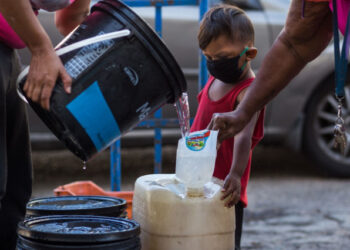A child wears a face mask as a preventive measure against the global COVID-19 coronavirus pandemic as his caretaker collects water in Caracas, on March 27, 2020. - Venezuela is facing the novel coronavirus pandemic while suffering a major gasoline shortage and with the country's water system collapsed, which has left many homes without running water. (Photo by Cristian Hernandez / AFP)