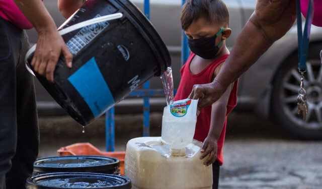 A child wears a face mask as a preventive measure against the global COVID-19 coronavirus pandemic as his caretaker collects water in Caracas, on March 27, 2020. - Venezuela is facing the novel coronavirus pandemic while suffering a major gasoline shortage and with the country's water system collapsed, which has left many homes without running water. (Photo by Cristian Hernandez / AFP)