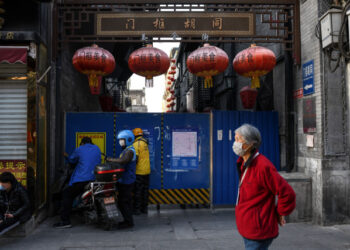 A woman (R) wearing a facemask as a preventive measure against the COVID-19 coronavirus walks past delivery riders waiting to pass food through a hole of a barrier in an alley in Beijing on April 2, 2020. (Photo by GREG BAKER / AFP)