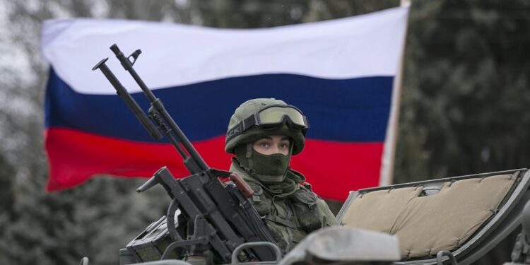 A pro-Russian man (not seen) holds a Russian flag behind an armed servicemen on top of a Russian army vehicle outside a Ukrainian border guard post in the Crimean town of Balaclava March 1, 2014. Ukraine accused Russia on Saturday of sending thousands of extra troops to Crimea and placed its military in the area on high alert as the Black Sea peninsula appeared to slip beyond Kiev's control. REUTERS/Baz Ratner (UKRAINE - Tags: MILITARY POLITICS CIVIL UNREST) - RTR3FVD5