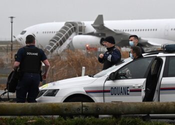 French customs officers stand next to a customs car with an Airbus A340 in the background which was grounded on the tarmac since December 21 over suspected "human trafficking", at the Vatry airport, north-eastern France on December 25, 2023. Two Indians passengers on the flight grounded since December 21, in the Marne region of France on suspicion of human trafficking will be presented to an examining magistrate on December 25, with possible indictment, while most of the other passengers are due to take off for India later on December 25. (Photo by FRANCOIS NASCIMBENI / AFP)