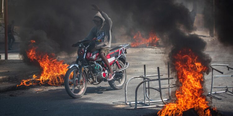 AME4424. PUERTO PRÍNCIPE (HAITÍ), 01/03/2024.- Una persona se transporta en motocicleta con incendios alrededor, cuando la tensión volvió este viernes a Puerto Príncipe con tiroteos entre la Policía y las bandas armadas, después de que la capital haitiana amaneciera en aparente calma tras la violenta jornada de la víspera con al menos cinco muertos y una veintena de heridos, este viernes en Puerto Príncipe (Haití). Según el último balance del Sindicato de Policía de Haití, se elevan a cinco los efectivos que murieron el jueves y cuyos cuerpos aún no han podido ser recuperados. En un principio, se informó de que cuatro policías perdieron la vida al atacar las bandas armadas una comisaría en Bon Repos, en Canaan, en el norte de la capital. EFE/ Johnson Sabin