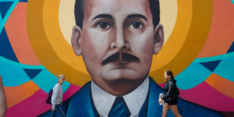 AME3403. CARACAS (VENEZUELA), 25/02/2025.- Personas caminan frente a un mural del beato José Gregorio Hernández este martes, en Caracas (Venezuela). El obispo auxiliar de Caracas, monseñor Carlos Márquez, expresó este martes su esperanza de que la canonización del beato venezolano José Gregorio Hernández, aprobada por el papa Francisco, sea una oportunidad para la construcción de una Venezuela unida y en paz. EFE/ Ronald Peña R.